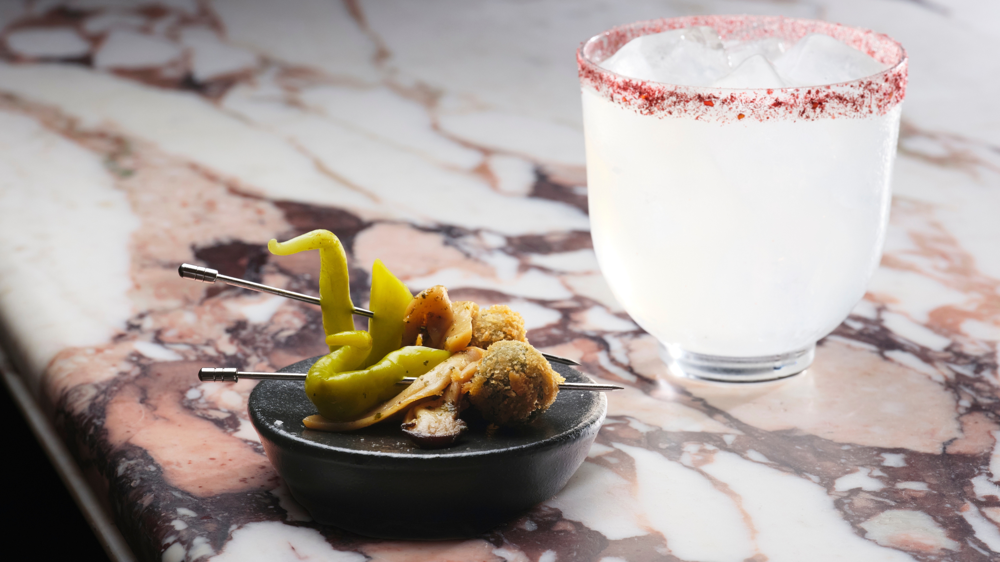 A purple calacatta marble tabletop holds a glass filled with a transparent cocktail whose rim has been covered in rose-tinted salt and a ceramic snack plate filled with colorful gildas placed on chrome sticks.