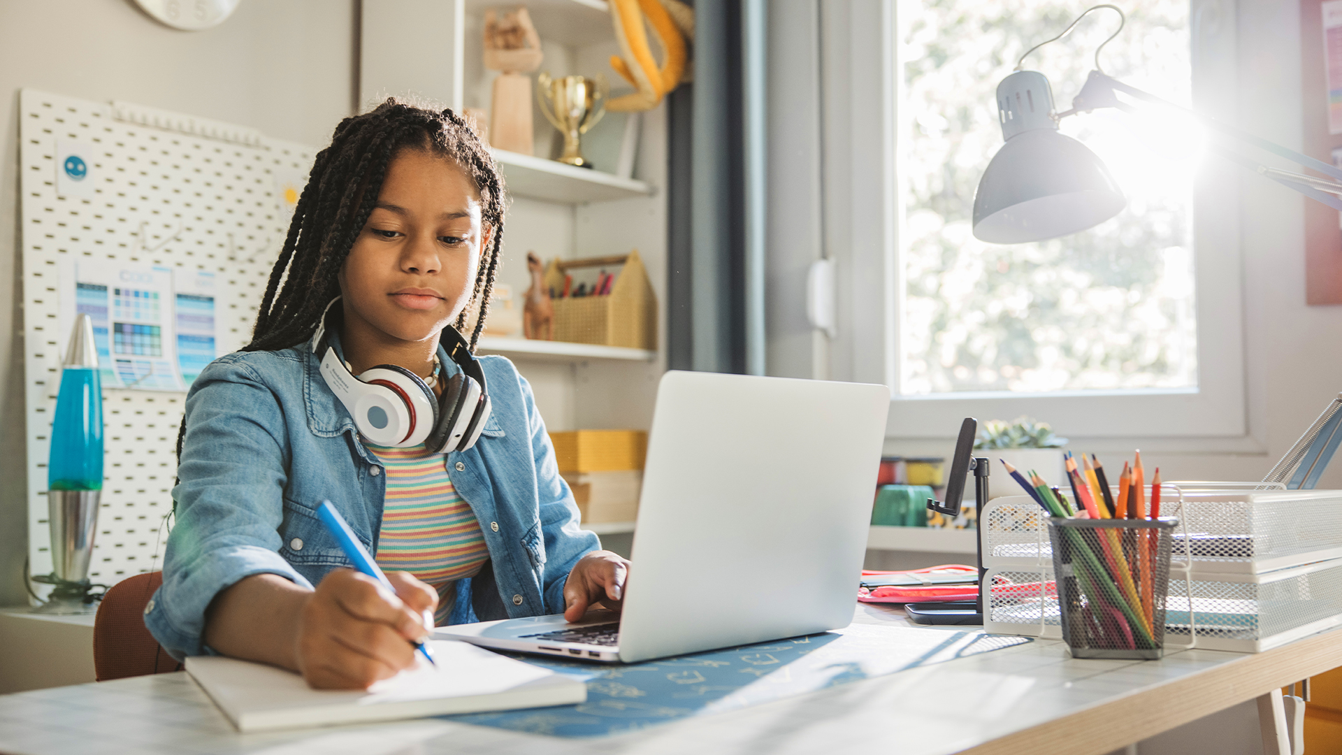 A young girl is doing homework, writing in her notepad while also working on her laptop