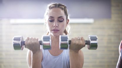 Fit young woman lifting two dumbbells up to shoulder height in a gym