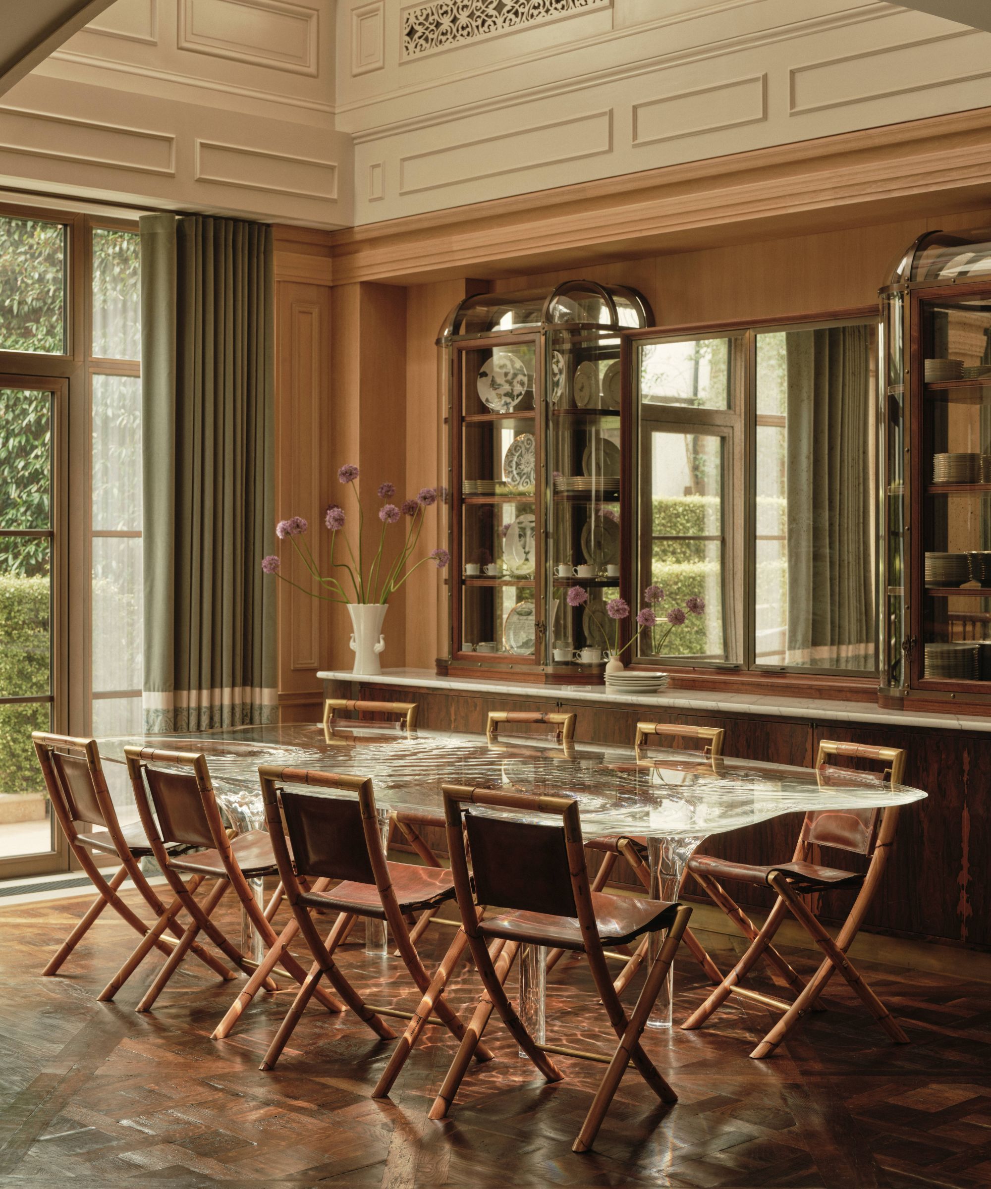 Dining room with a modern clear glass table and wooden chairs. A modern cabinet is behind with a mirror and French doors with green drapes let in light