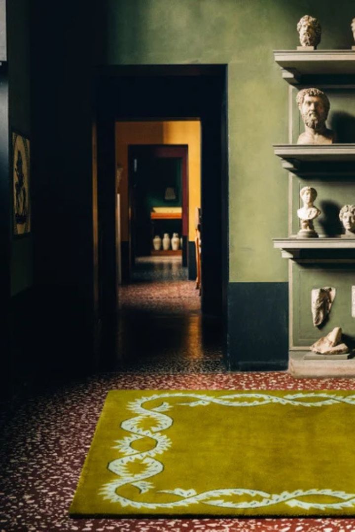 A specled terrazzo floor in red and white marble is topped by a tufted rug in olive green and surrounded by marble busts in a spacious palazzo room.