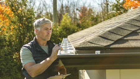 A mature man stands on a ladder to inspect the roof of his house.