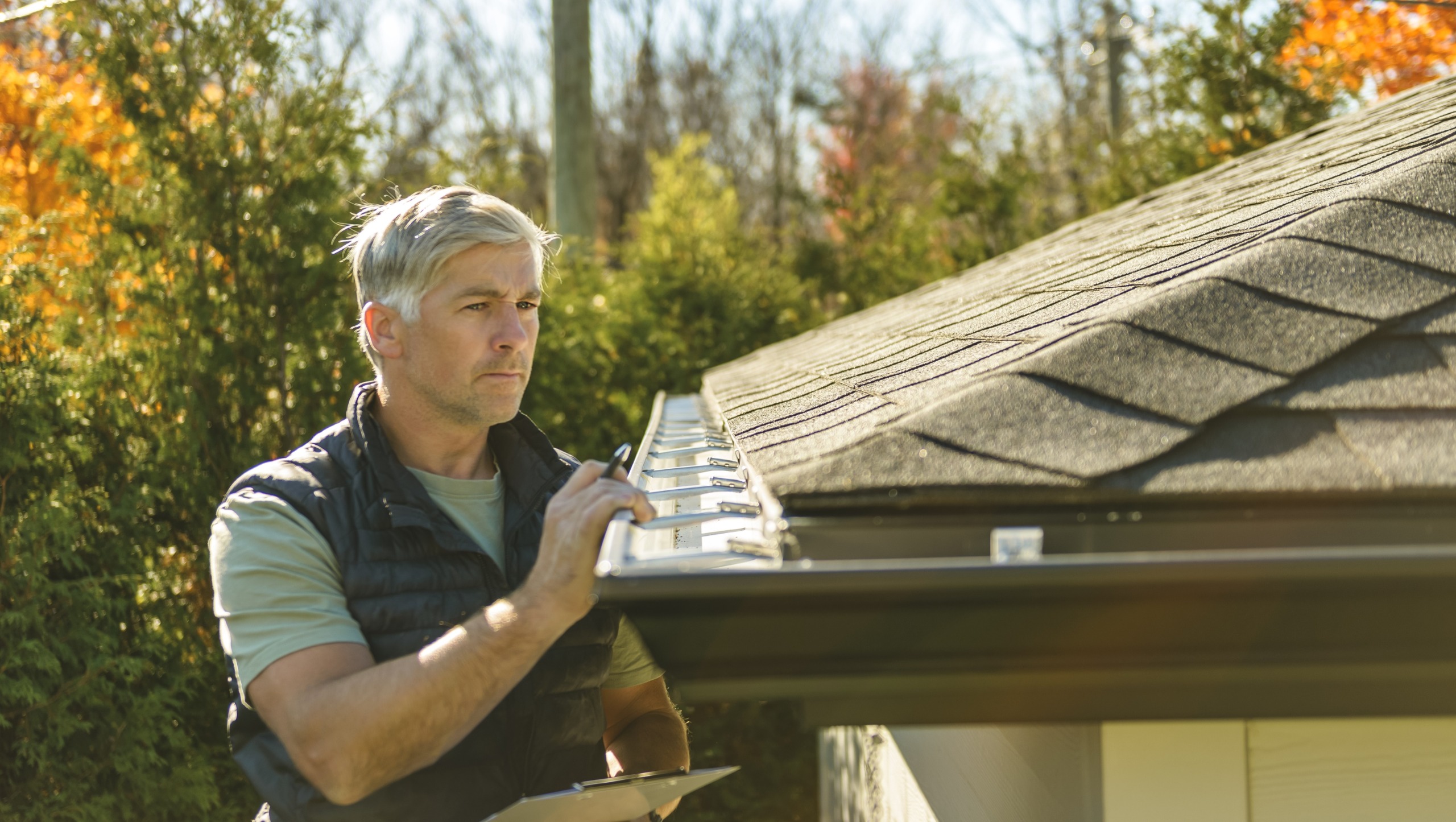A mature man stands on a ladder to inspect the roof of his house.