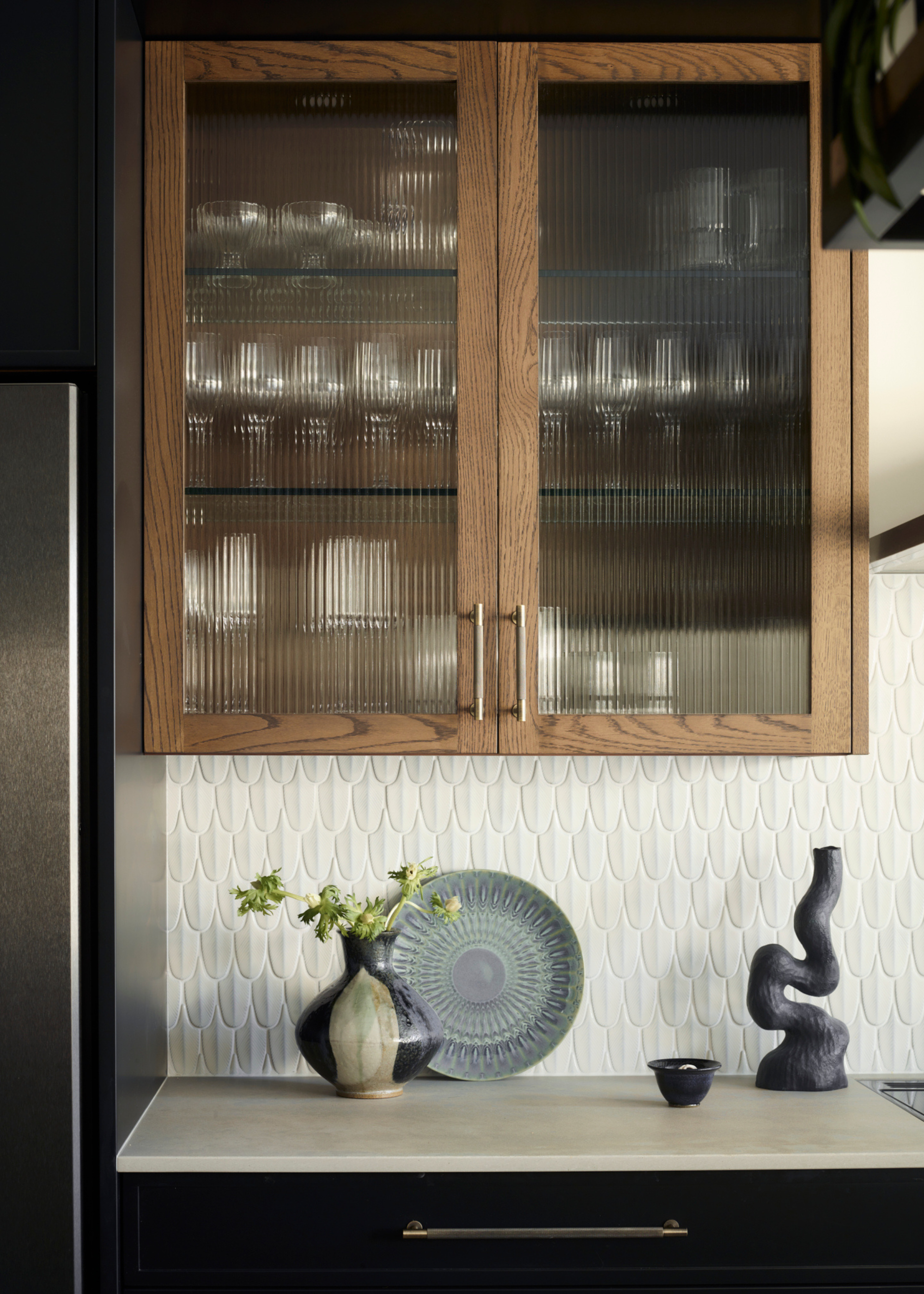 A modern kitchen with wooden cabinetry and fluted glass fronts and a scalloped white tile as the backsplash