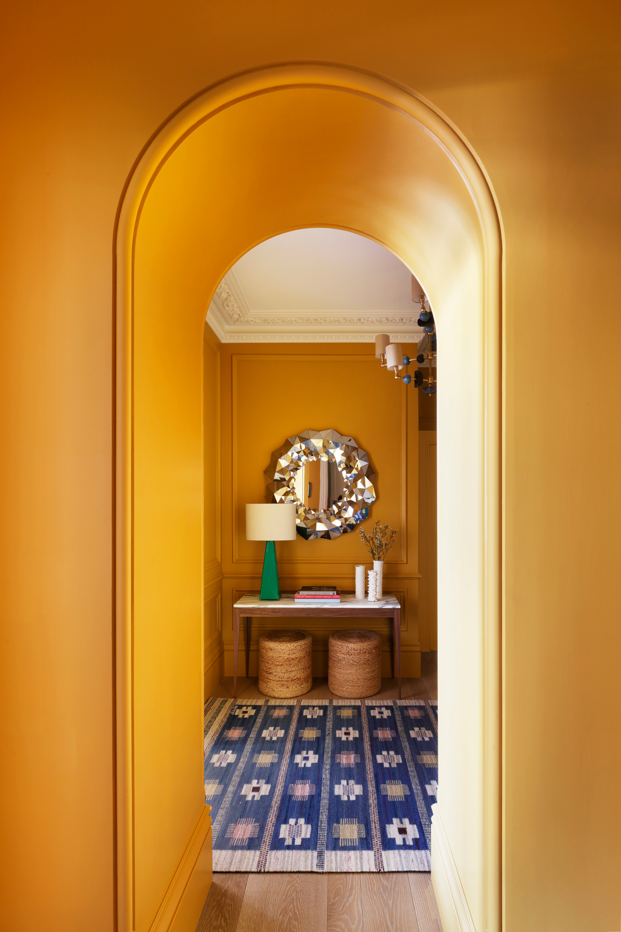 View to hallway through arched doorway with mustard walls, blue and white rug and wood and marble console table with chrome mirror above