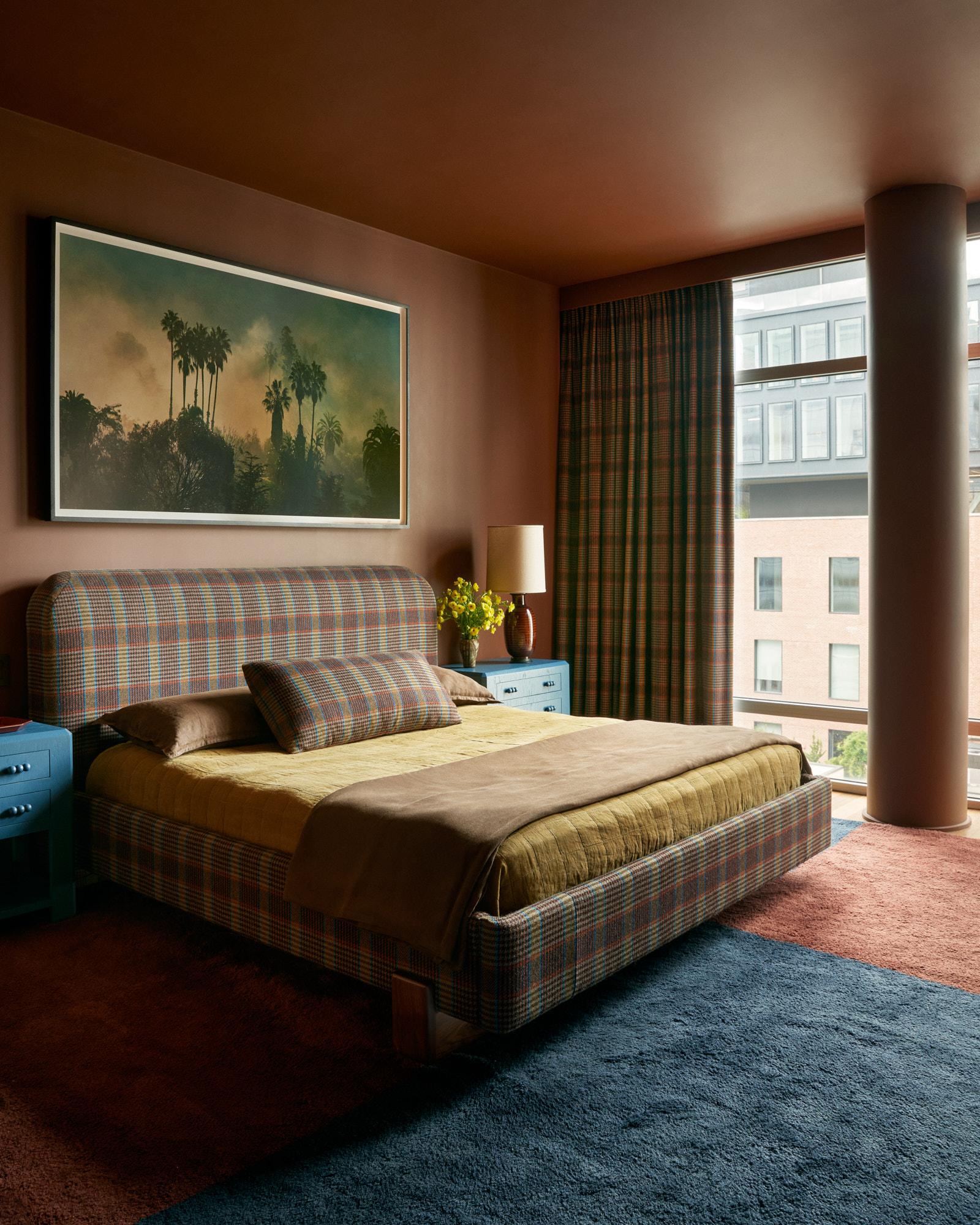 A modern bedroom with plaid curtains, a plaid bed frame and a matching plaid bolster pillow with rust walls and ceiling and a portion of carpet together with dark blue that matches the nightstands