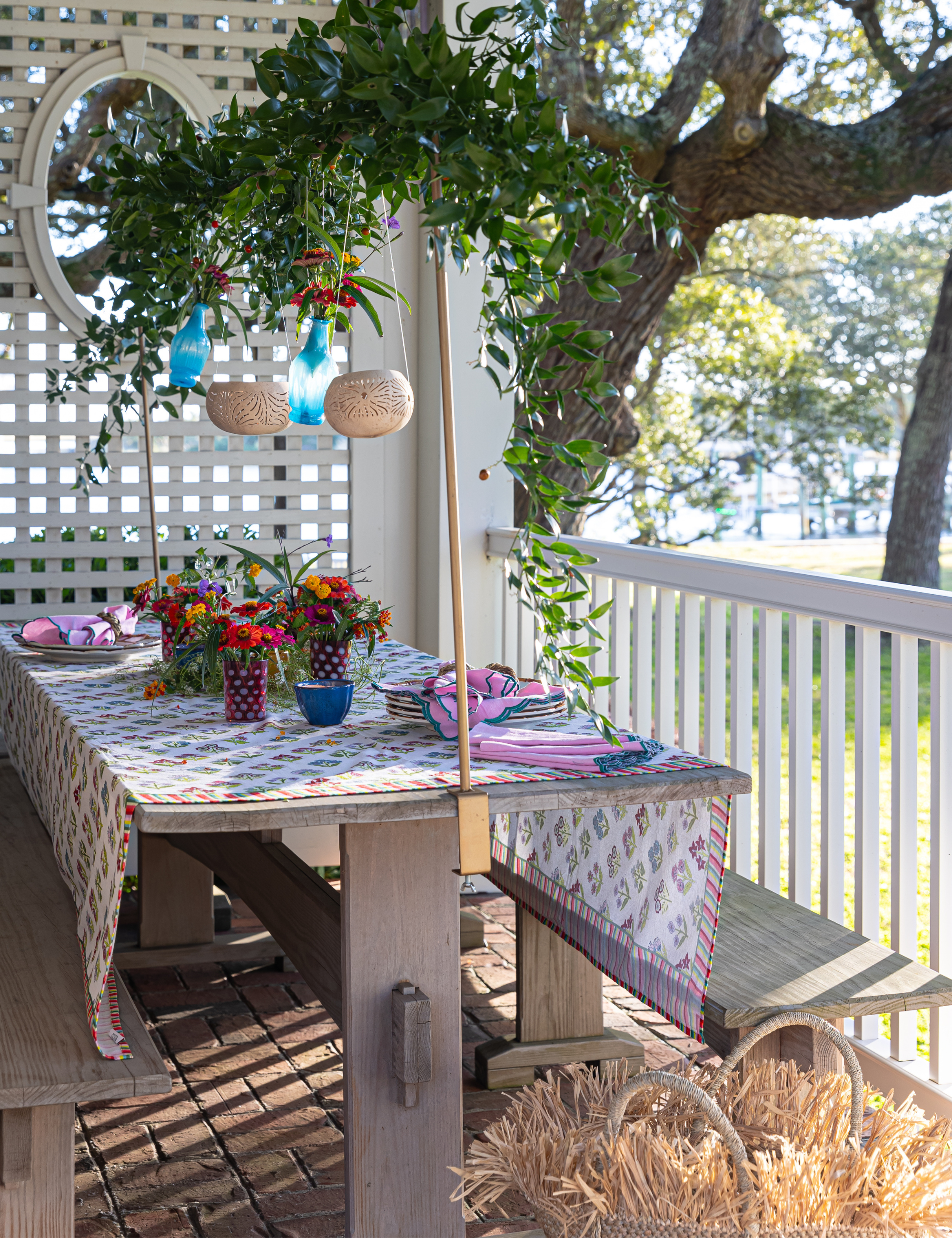 Outdoor dining area featuring a rustic picnic table styled with a floral tablecloth, dinnerware, and bohemian lanterns suspended from a tree branch overhead.