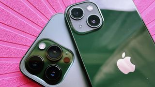 A green iPhone 13 resting atop a green iPhone 13 Pro, on a bright pink background