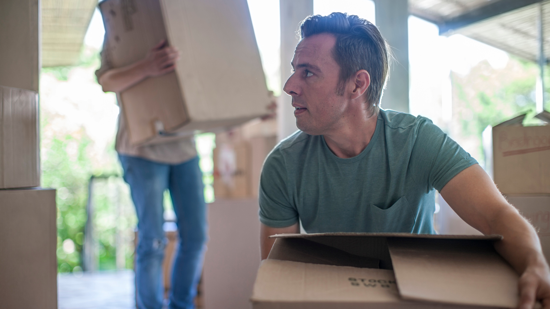 Man crouched down holding a cardboard box, with a person carrying a large cardbox in the background