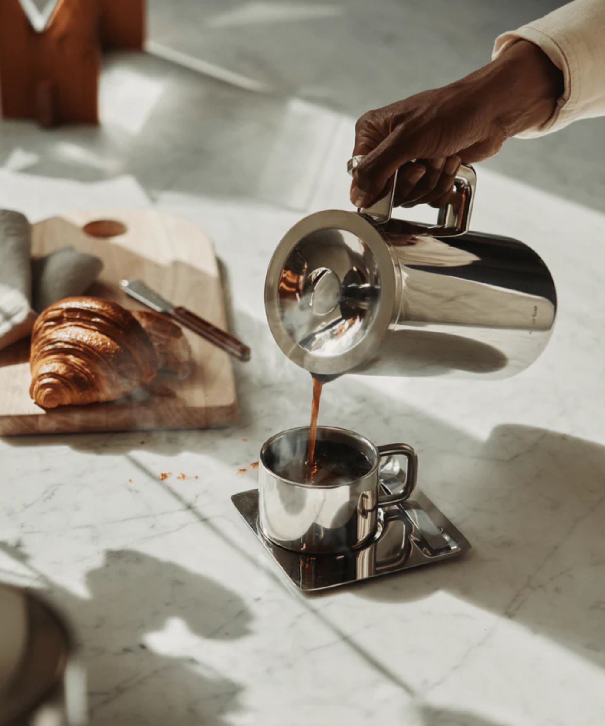 The stainless steel aarke Coffee press being used to pour steaming coffee into a stainless steel cup on a marble kitchen counter. There is a croissant on a cutting board behind it.