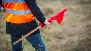 back of male wearing orange hi vis, blue jacket and jeans stood on bare land holding rolled up red flag