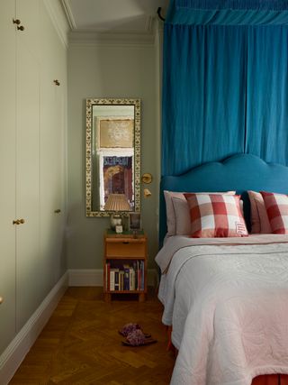 A pale green bedroom with a bed that has a bright blue canopy/drapery behind it. The pillows on the bed are red and white gingham, and the duvet is white.