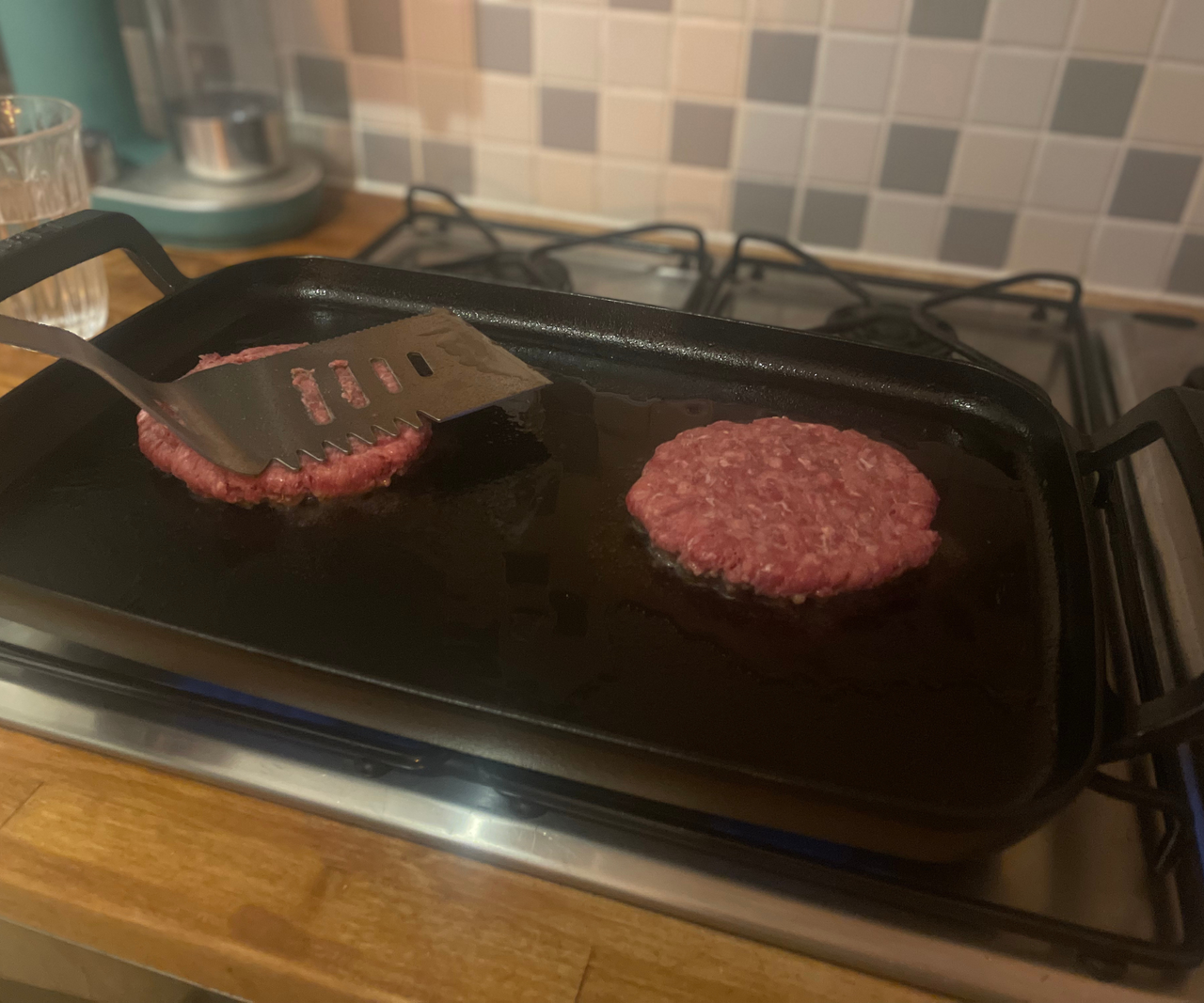 Two raw burger patties being fried on a flat griddle pan across to gas burners.