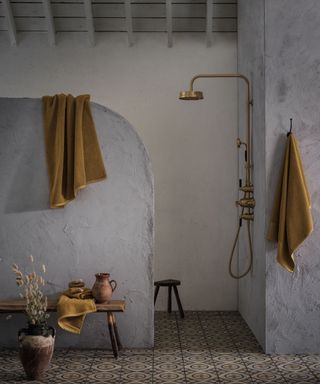 A wet room with a partition wall with exposed walls. Patterned floor tiles and a gold shower head. A mustard coloured towel hangs outside the wet room on a hook and a small wooden bench with a terracotta jug on top.