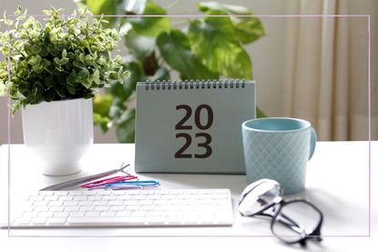 A 2023 calendar on a desk with a plant, pair of glasses, keyboard and a mug