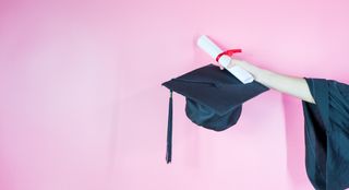 A hand holding graduation cap and diploma on pink background
