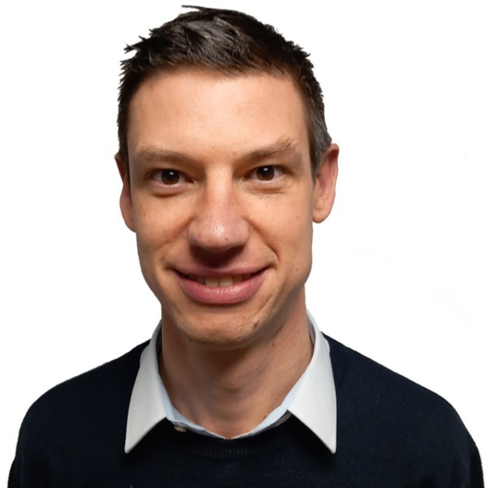 Close up headshot of a smiling man with short brown hair wearing a white shirt and black jumper