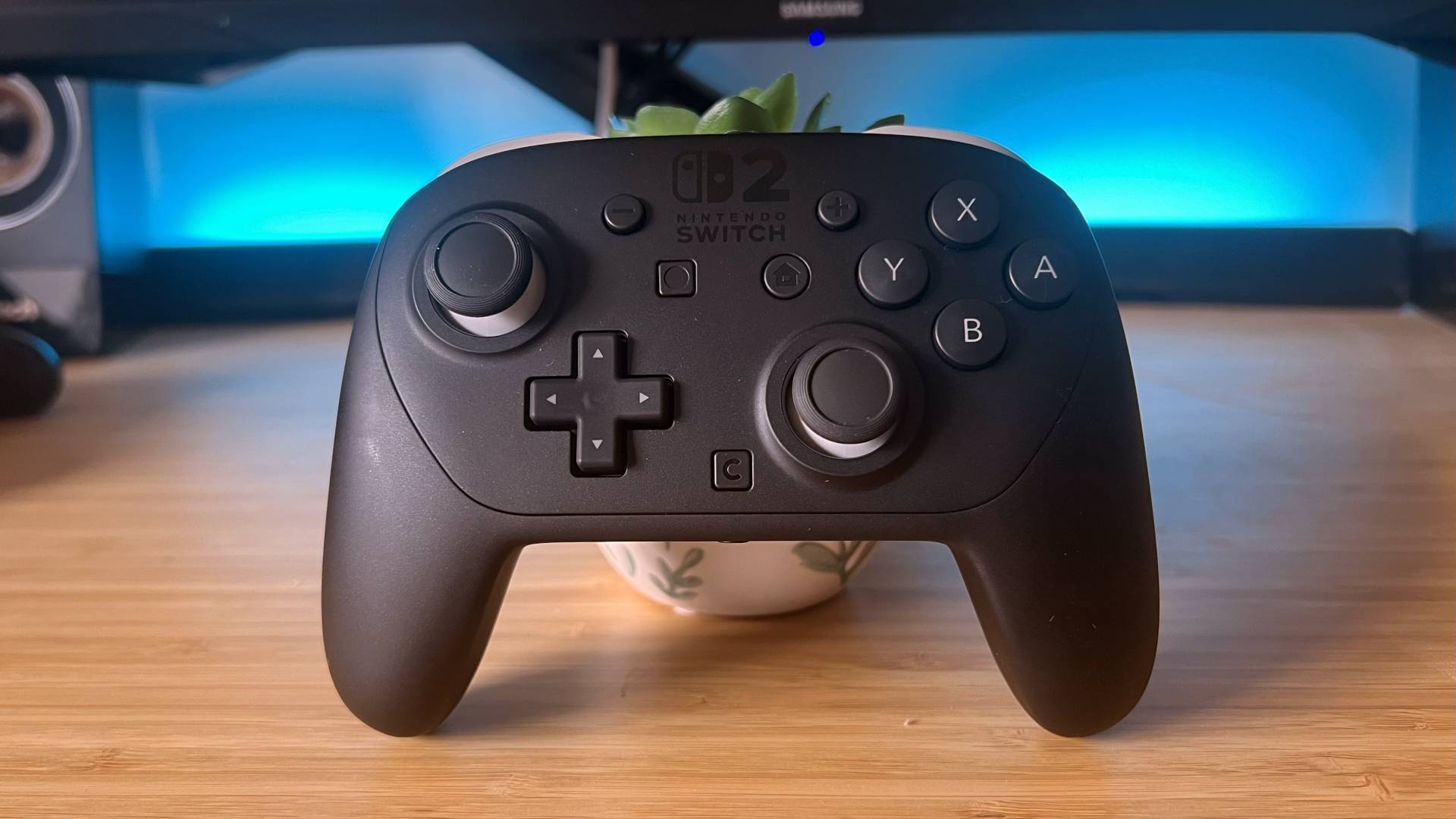 Switch 2 Pro Controller leaning against small plant on woodgrain desk.