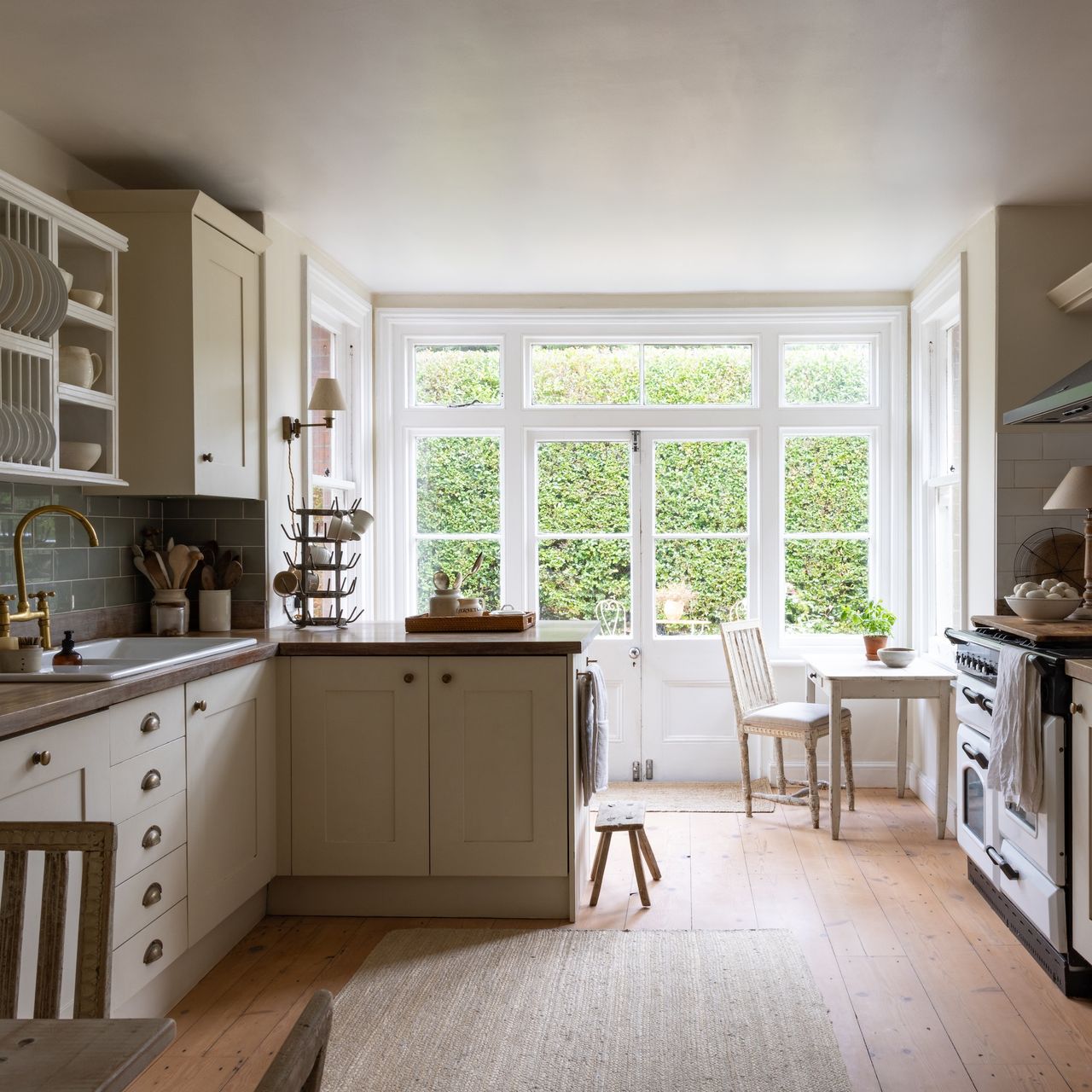 country-style kitchen with cream shaker units and range cooker, wooden floor and large windows at end of room