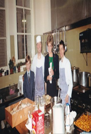 Prince William and Princess Diana standing in a kitchen at charity The Passage with two chefs wearing aprons