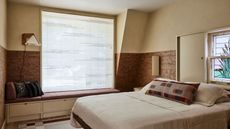 bedroom with window seat, blinds, cork wall wrap and plaster ceiling, mirror behind the bed, and cream bedding with patterned cushion
