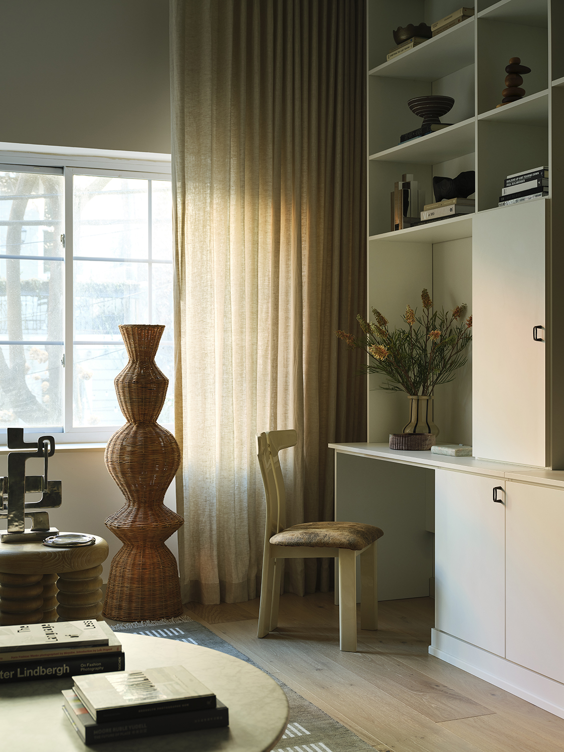 Image of a room with a window with tan curtains and a large, white built-in with flowers and other decor on the shelves.