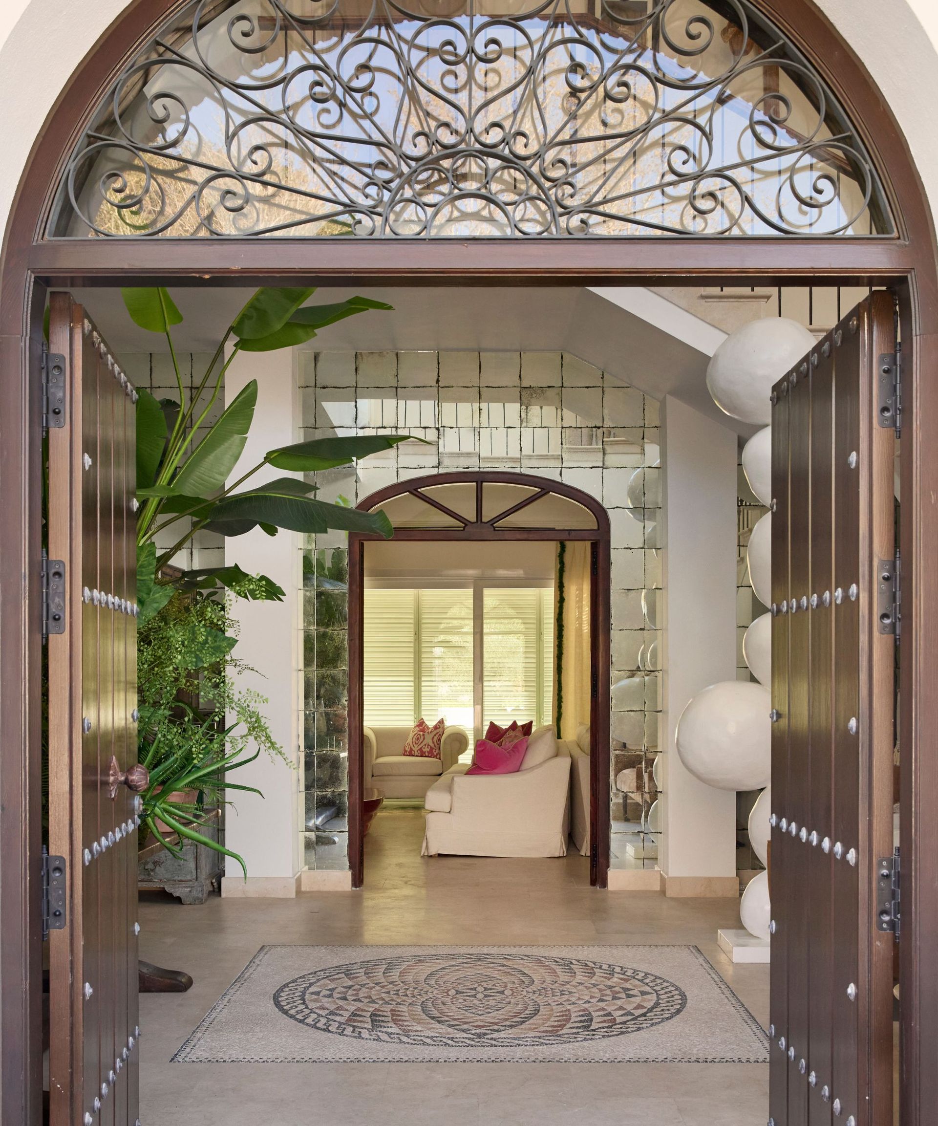 Welcoming ornate double front door which is open and showing a large stone-floored hallway, with sight of a cozy living room with cream upholstered sofas in the background