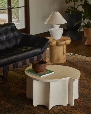 A modern rustic-furnished living room features a round coffee table composed of two half-moon parts, a leather and wood, modernist couch, a side table in wood, and a white lamp standing on top of it. Further decorations include a wide, orange-shaded rug and plants.