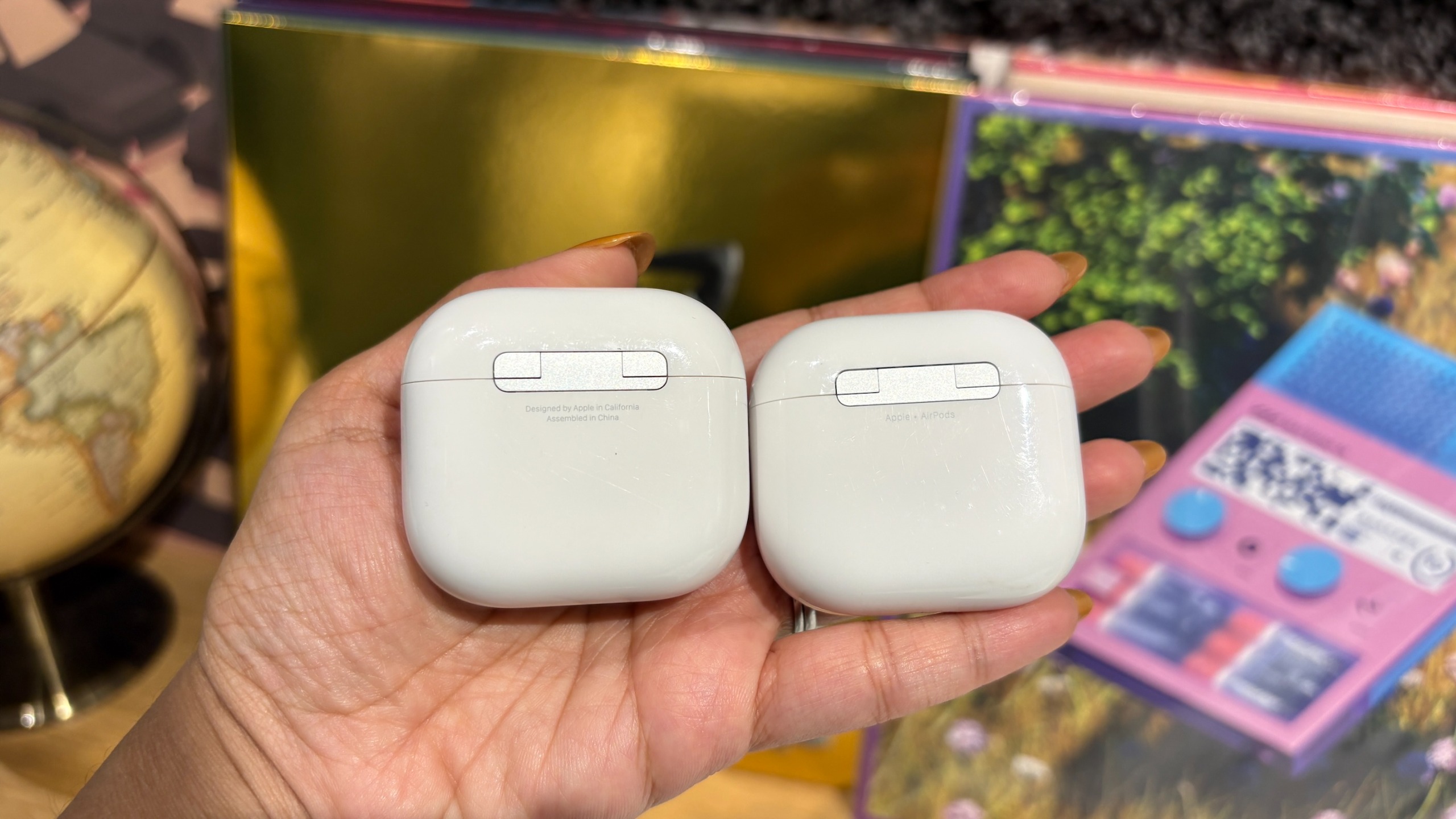 The rear of the AirPods 5 and AirPods 4 charging cases held side by side in a hand.