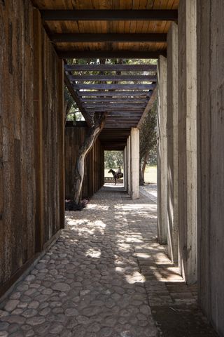 A pathway that surrounds the house, is covered with wooden beams and boards. The floor is made out of small stones. In the distance, we see a man riding a horse.