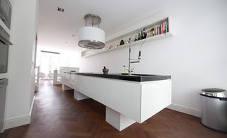 A kitchen space featuring white and silver decor with dark wood flooring. A long island with black marble top and marble pistle & mortar. Long white walled shelf with items placed in the shelf