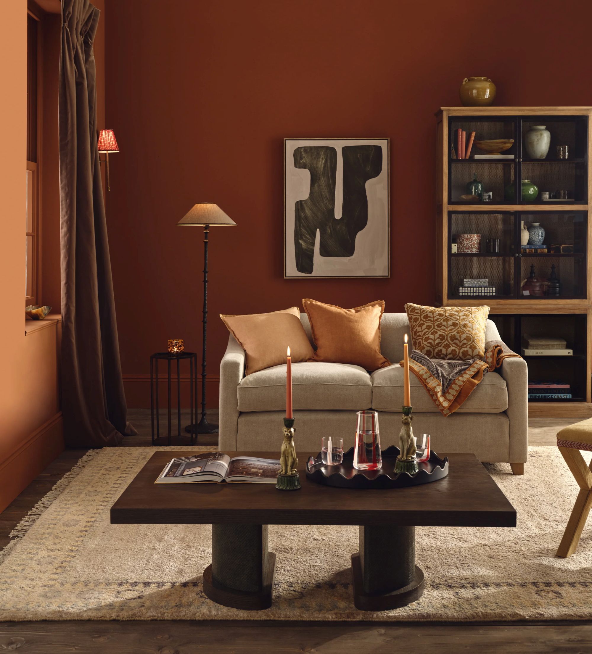 a living room with burnt orange walls, a small two seater couch in light tan, a brown coffee table with candle holders and a serving tray, and a light tan rug with an abstract floral motif along its border