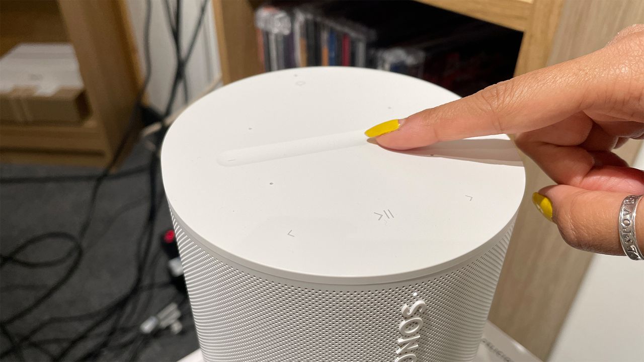The top panel of a white Sonos Era 100 smart speaker. A person with yellow nail varnish has placed their finger on top.