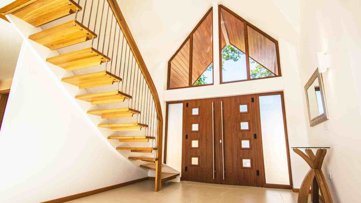 A large hallway with vaulted ceiling and large double timber front door and glazed gable above. The hallway, painted in white, features a timber and steel floating staircase, by Complete Stair Systems, to one side.