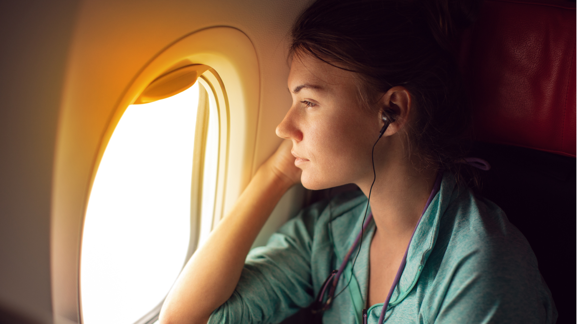 A woman sits on an airplane wearing corded, in-ear headphones while looking out the cabin window.
