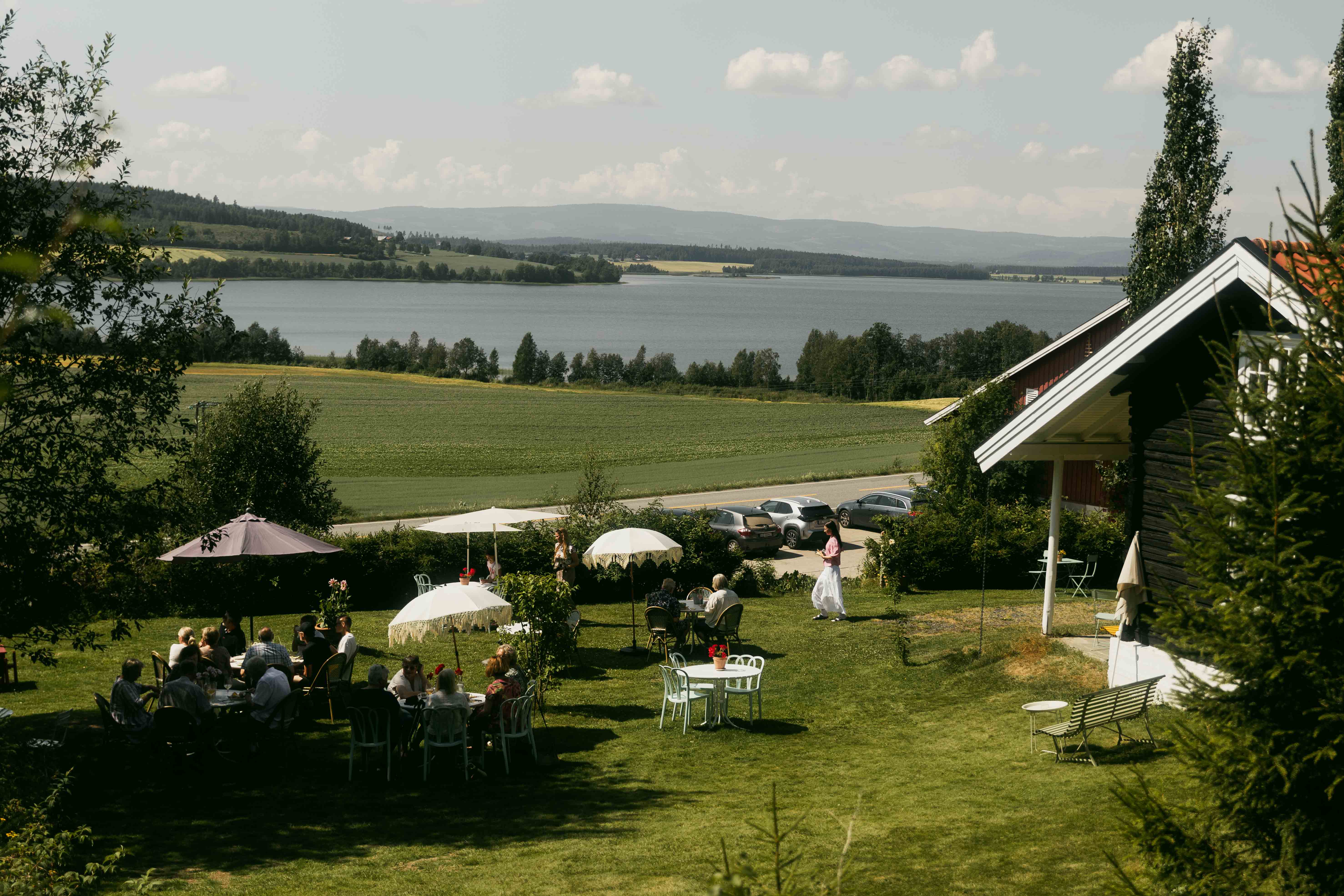 A bucolic landscape, showing verdant vegetation and a lake, is decorated with retro-inspired parasols as a crowd of people enjoys their time outdoors. 