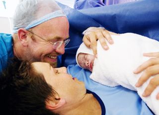 A woman in a hospital bed, smiling and holding her newborn baby after having given birth via C-section. A man is crouched at her head, smiling and looking at the baby.