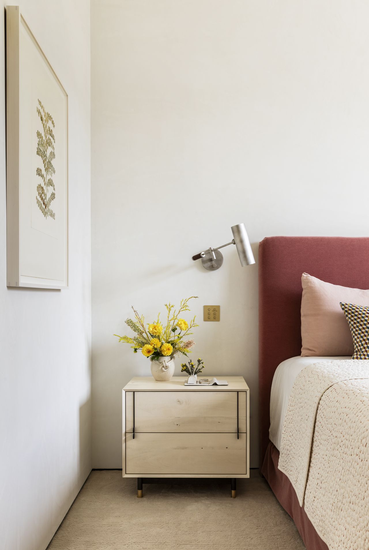 A view of a bedside table beneath framed art alongside a maroon headboard