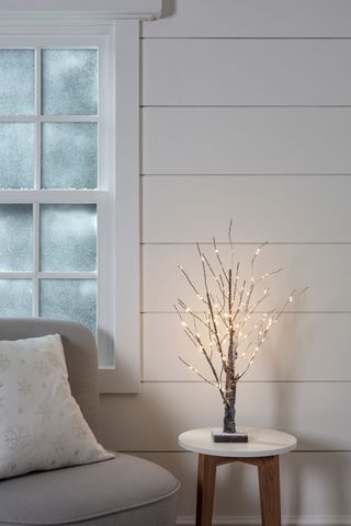 A frosted, snow-effect pre-lit battery twig tree with white shiplap decor and grey chair