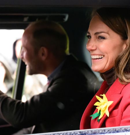 Prince William riding in a car waving to fans out a window sitting next to Princess Kate, wearing a red coat with a yellow daffodil pin and laughing