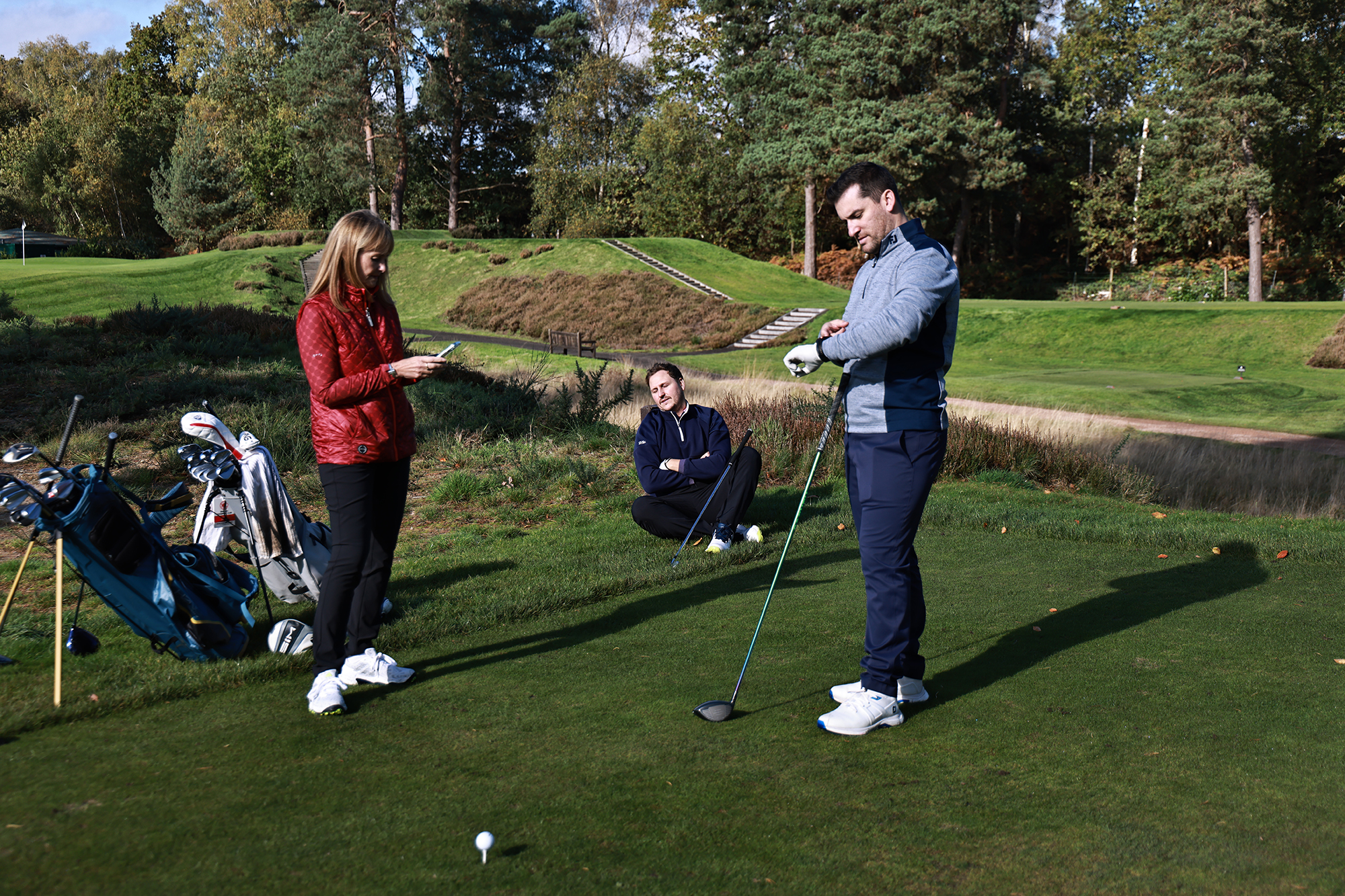 Three golfers waiting on the tee due to slow play ahead of them, with one sitting on the floor, one checking their phone and one looking at their watch
