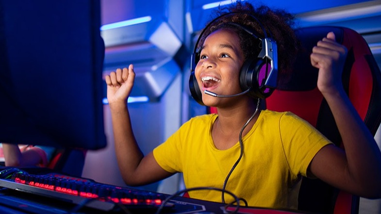 A smiling young girl playing a game on a computer with a headset on.