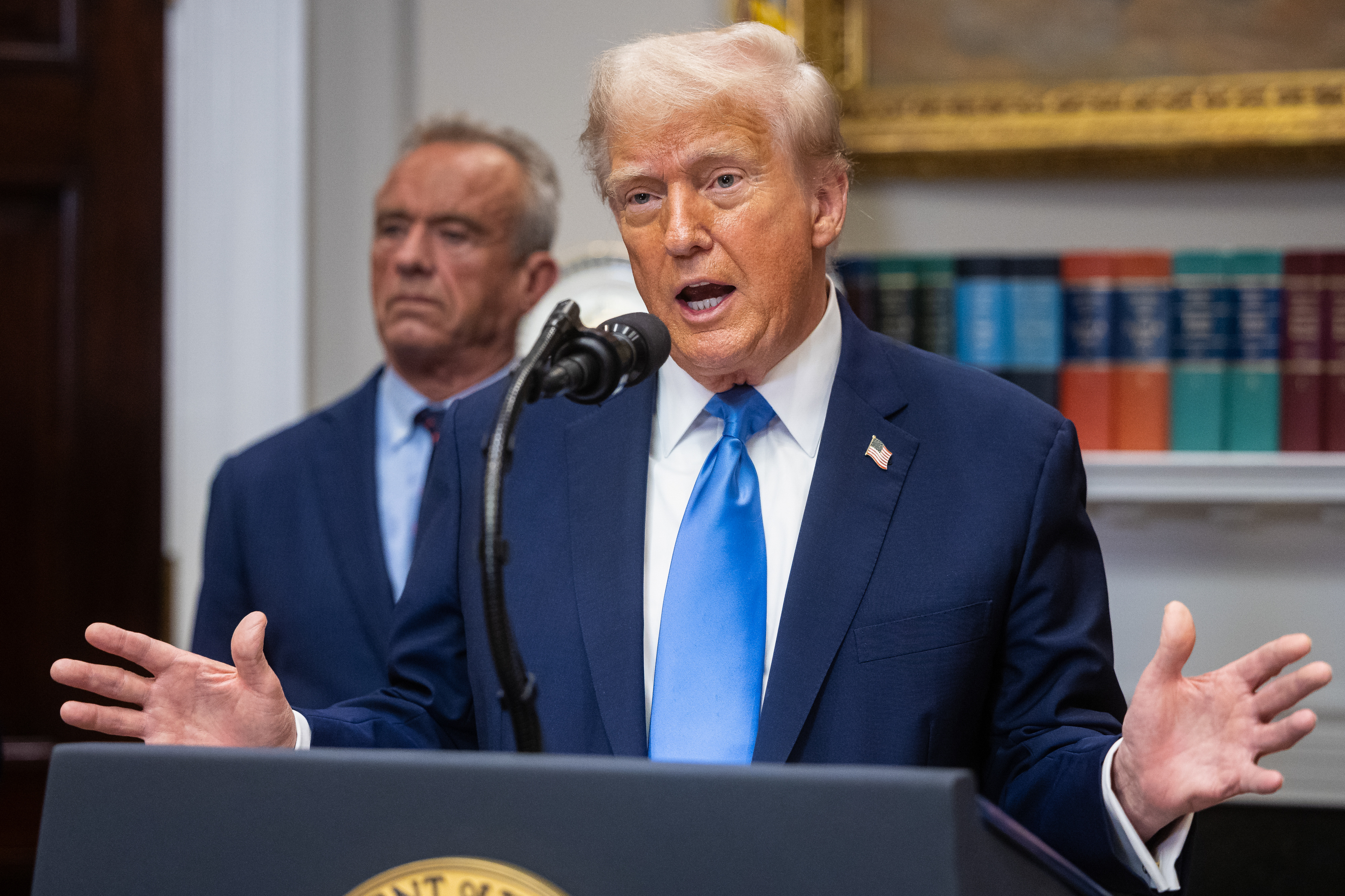 Trump at a podium talks about autism with RFK Jr in the background.