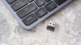 a dongle of a black and grey budget wireless Bluetooth keyboard with a full-size layout and a numerical keypad is photographed against a blue background