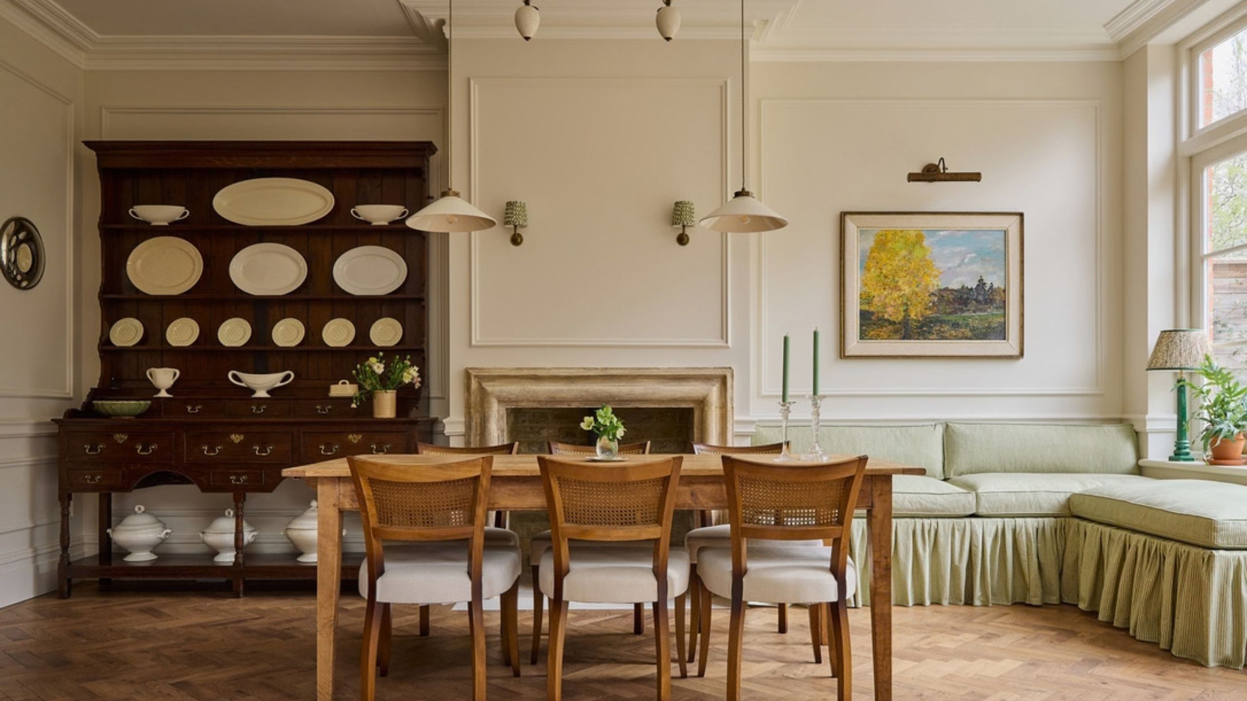 Open dining room with green skirted banquette seating in the corner, a long wooden dining table with rattan dining chairs, a fireplace at the center, and a dark mahogany dresser with white ceramics.