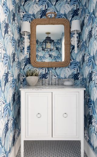 small powder room with blue leafy wallpaper, rope style mirror, white wall lights, vintage vanity with marble countertop, gray hex tile floor tiles