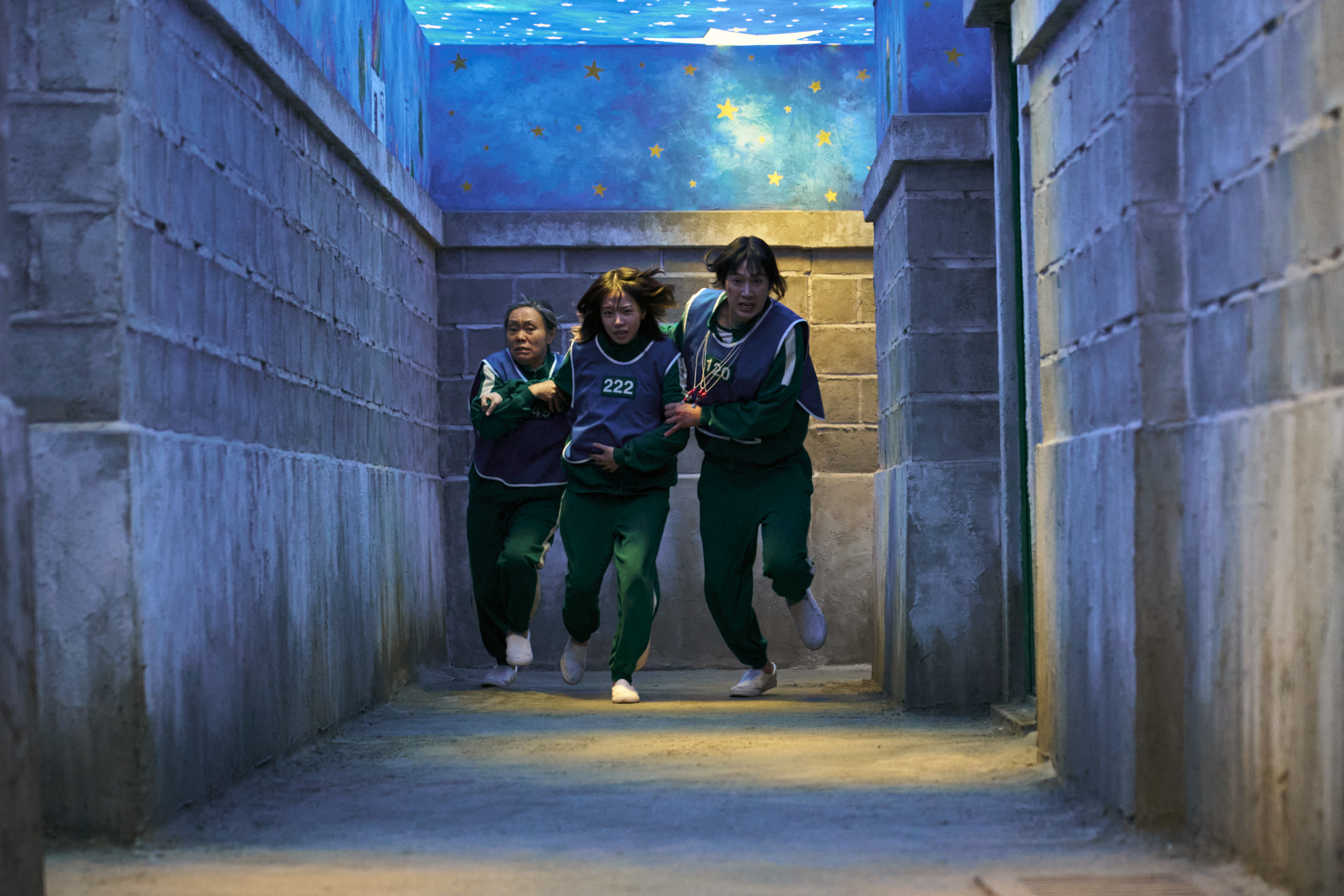 Three women (Kang Ae-sim as Geum-ja, Jo Yu-ri as Jun-hee, Park Sung-hoon as Hyun-ju) run down a bricked hallway with a roof of a painted starry night, in Squid Game S3.