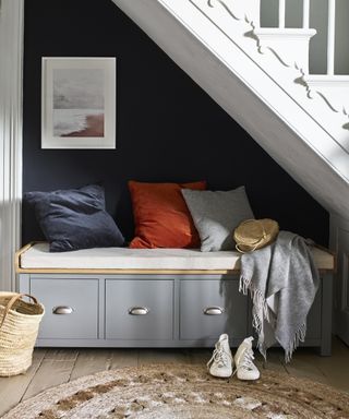 Modern entryway bench ideas with a grey shoe storage bench installed under a staircase with blue wall and blue and orange cushions