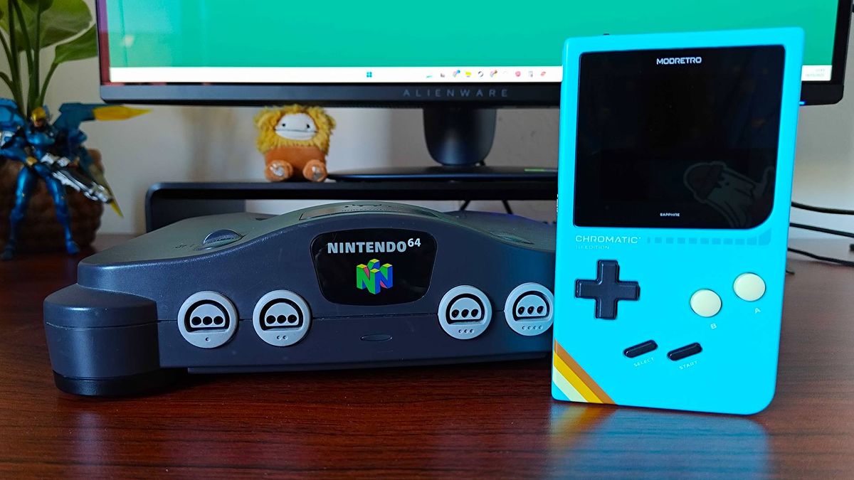 Nintendo 64 console next to blue Modretro Chromatic Game Boy handheld on woodgrain desk with Alienware monitor in backdrop next to plant.