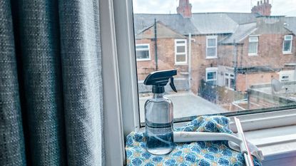 Learning how to clean windows is useful. Here is a windowsill with a glass cleaning bottle, a blue and white patterned cloth, and a squeegee on it, with a gray curtain panel in front of it and a window with brick houses beyond it